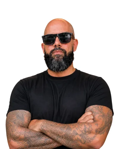 Allan Oliveira, consultor de SEO, de braços cruzados, camiseta preta e óculos escuros, em retrato da cintura para cima sobre fundo claro